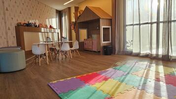 Um quarto espaçoso com uma grande janela, um respiradouro no teto, uma área de recreação com tapete de espuma colorido e uma cozinha de brinquedo infantil.