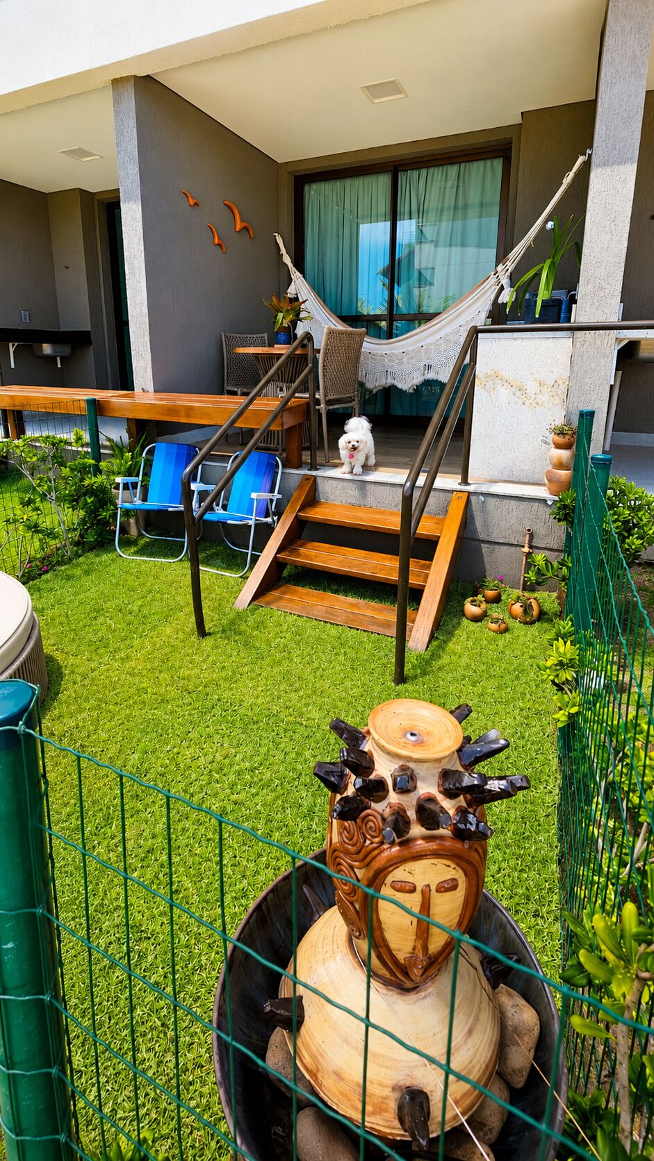 Un espacio exterior moderno con una hamaca, sillas azules, un perro pequeño y una maceta decorativa.