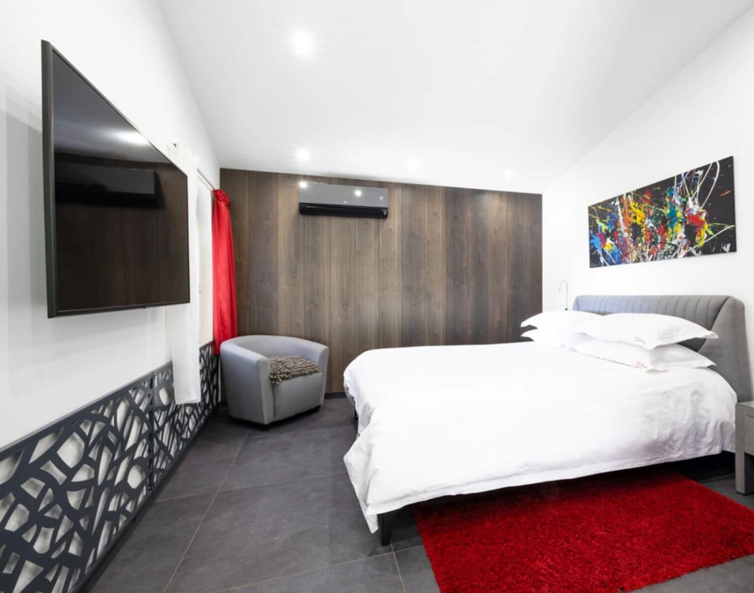 Quarto moderno com cama grande, TV de tela plana na parede, tapete vermelho decorativo e painel de madeira na parede.