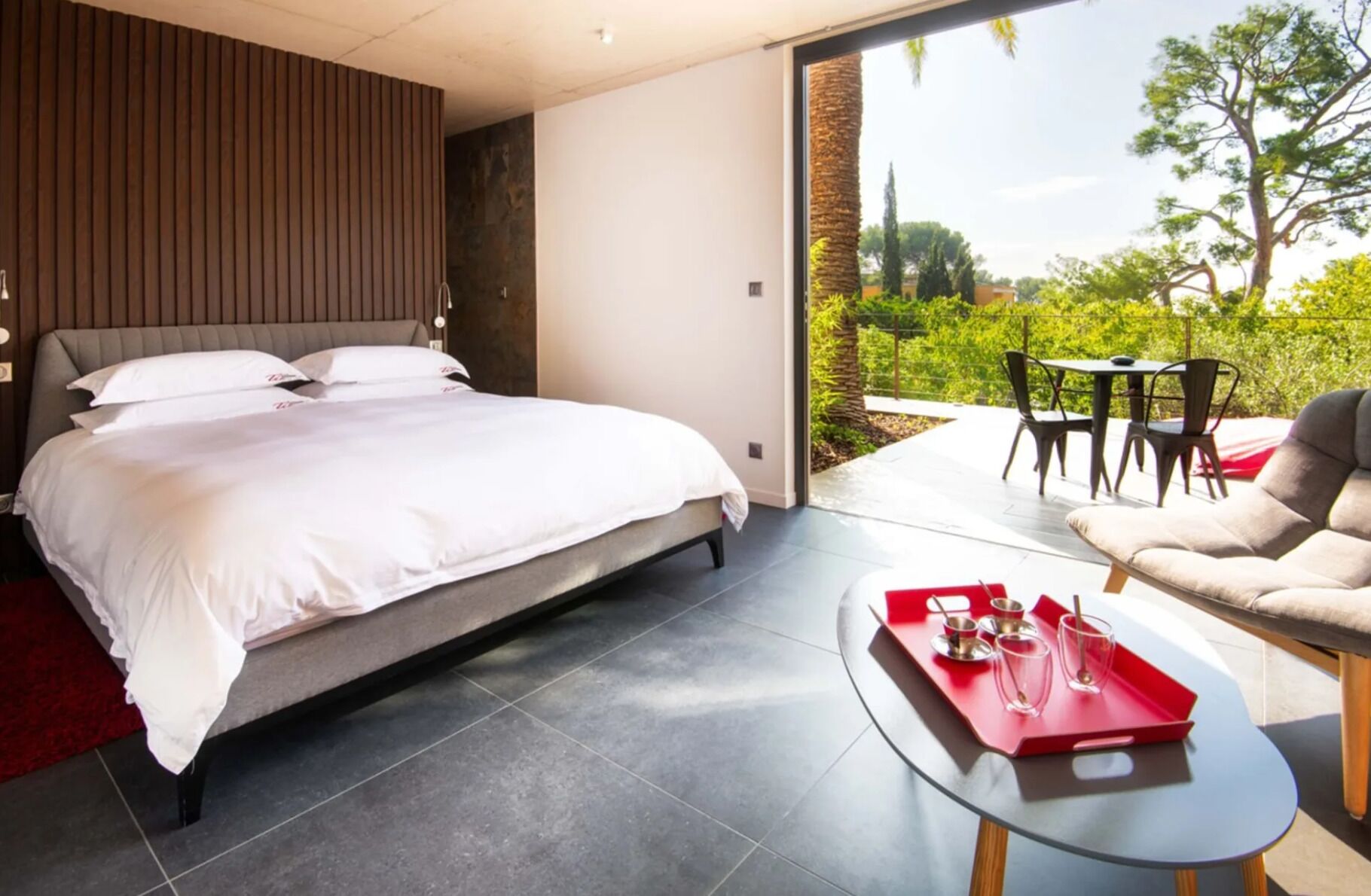 A modern bedroom with a large bed, a glass coffee table with red trays, and a view of the outdoors.