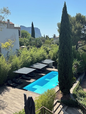 A swimming pool with lounge chairs and umbrellas, surrounded by greenery and a mountain in the background.