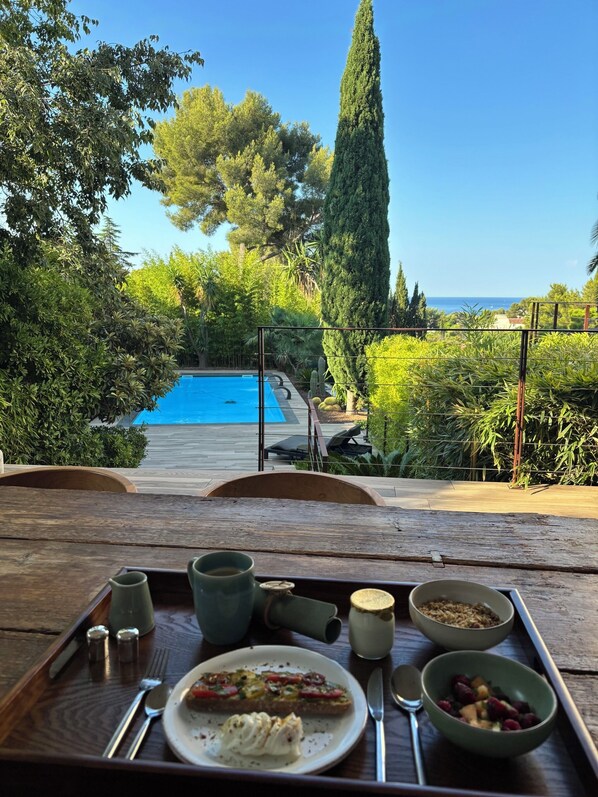 Mesa de comedor con una bandeja que contiene alimentos para el desayuno, con vista a una piscina y vegetación exuberante.