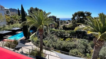 Un espace piscine offrant une vue dégagée sur la mer, entouré de palmiers et surmonté d’un auvent rouge.