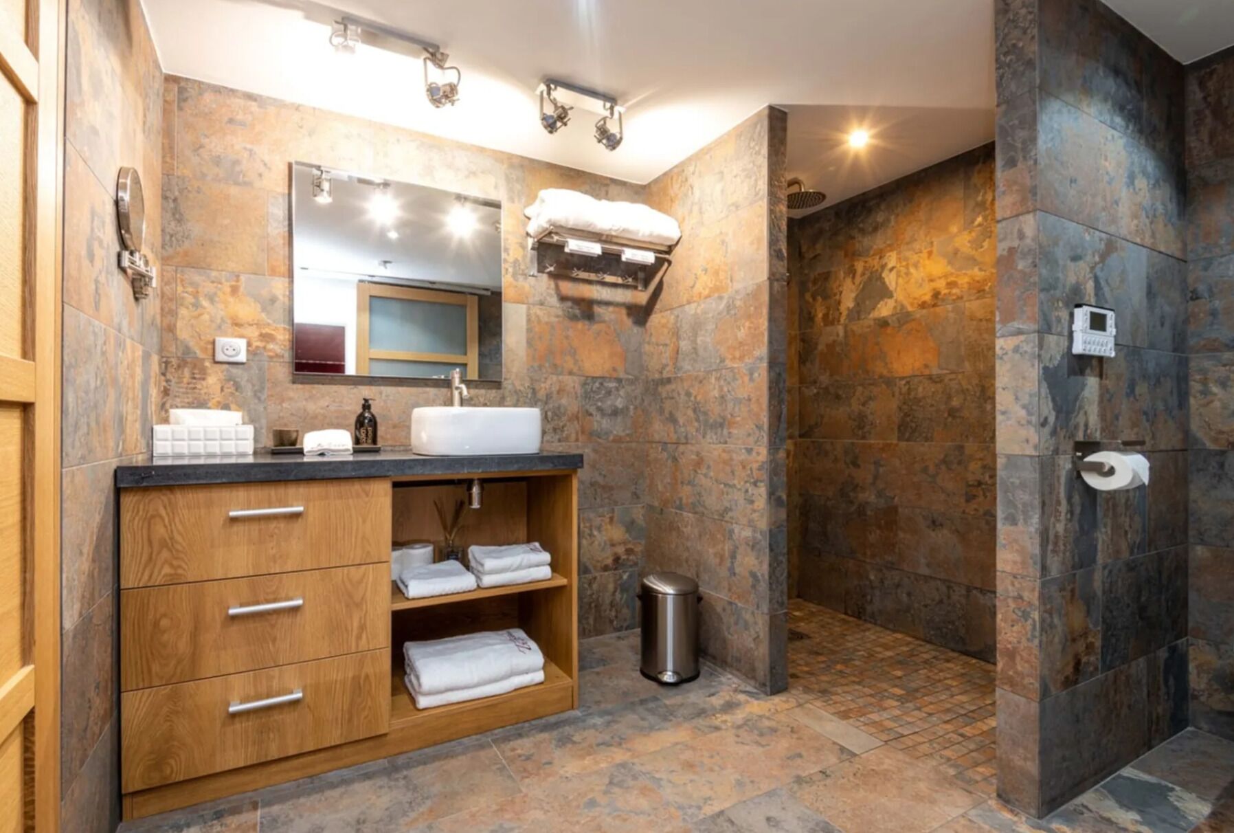 Un baño moderno con pared de piedra, tocador de madera, lavamanos y zona de ducha.