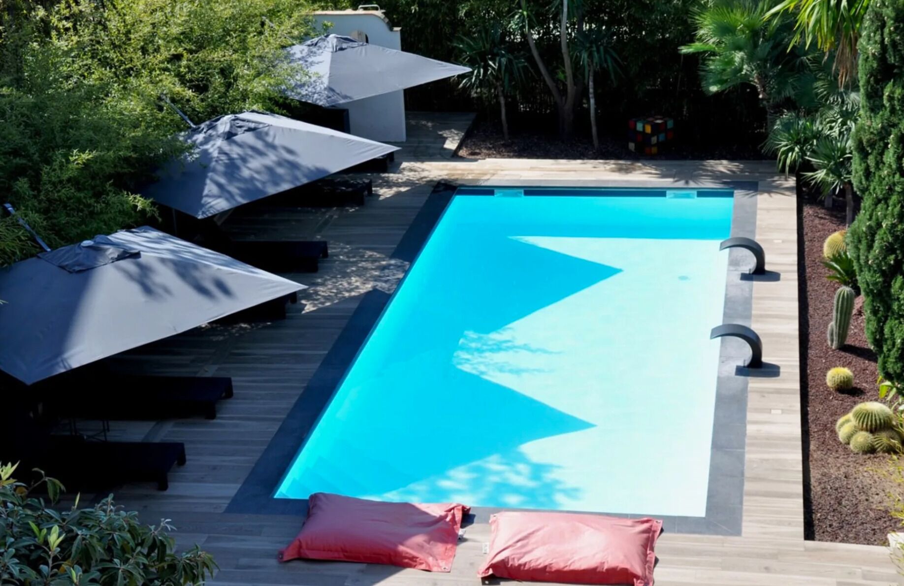 Una piscina rectangular con interior azul, rodeada por una terraza de madera. Hay dos sombrillas y dos sillas reclinables en la terraza.