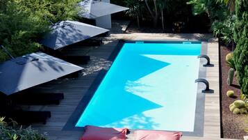 Una piscina rectangular con interior azul, rodeada por una terraza de madera. Hay dos sombrillas y dos sillas reclinables en la terraza.