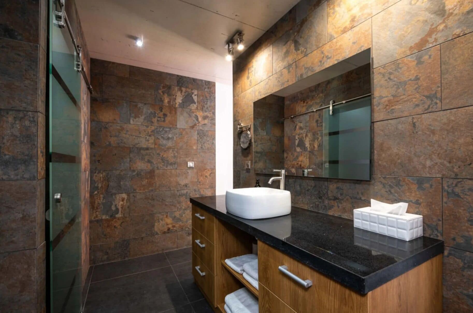 Une salle de bain moderne avec un mur en pierre, un grand miroir, un lavabo blanc et un meuble-lavabo en bois.