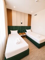 Hotellihuone, jossa on kaksi erillistä sänkyä, vihreäverhoiltu pääty ja puinen yöpöytä sekä seinään kiinnitetty valaisin.