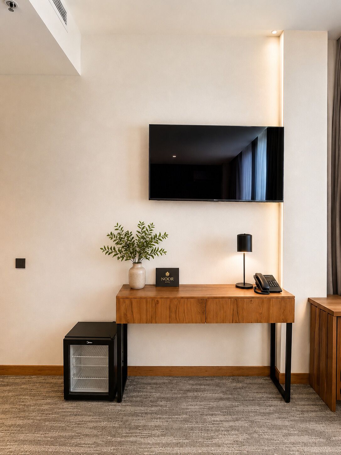 Quarto de hotel com televisão de tela plana na parede, mesa de apoio de madeira com uma planta, luminária e telefone.