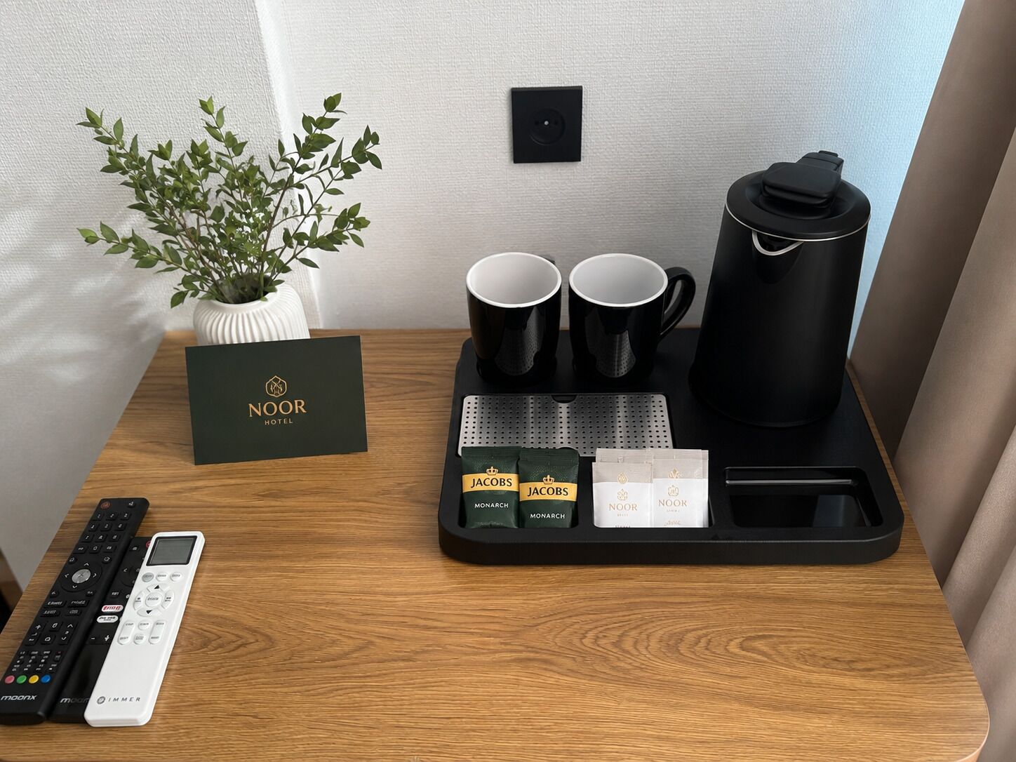 A hotel room with a wooden desk, a plant, a black tray with coffee cups, a water bottle, a remote control, and a card with the hotel name.