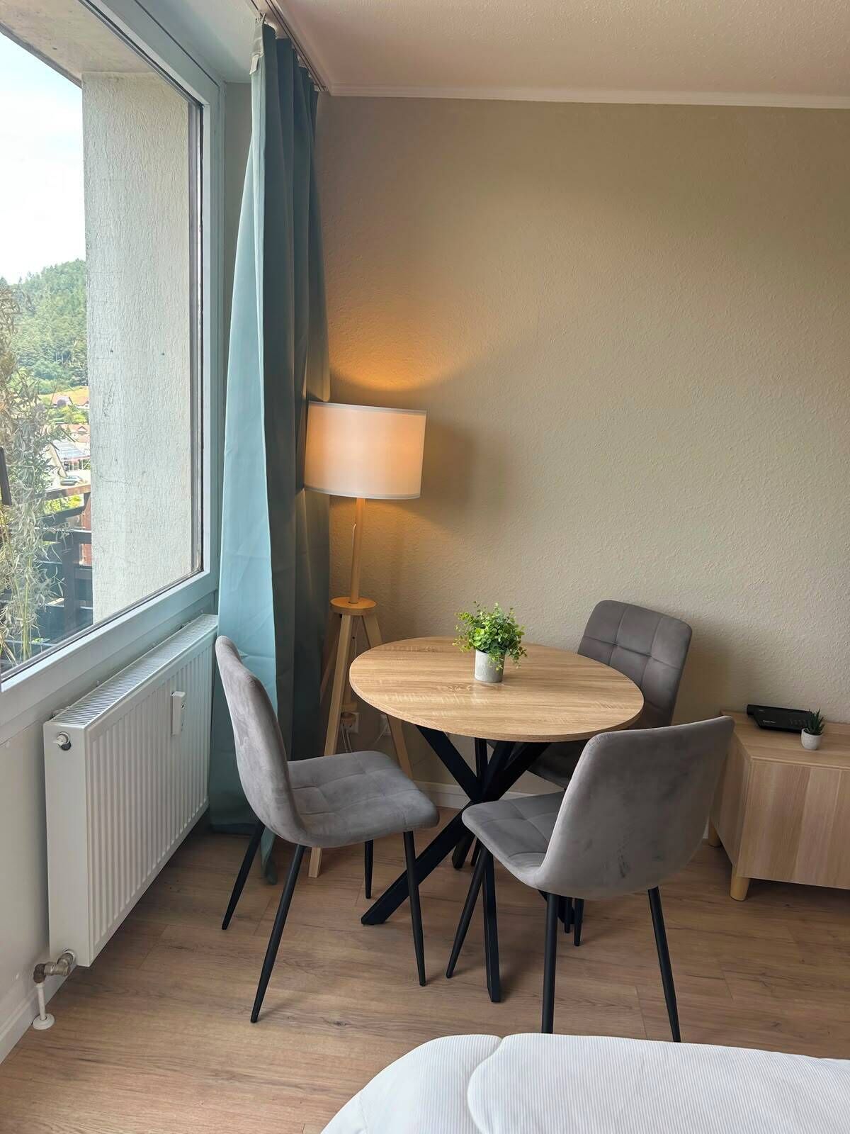 Ein Zimmer mit einem runden Holztisch, vier grauen Stühlen, einem kleinen Topfpflanzen, einer Lampe und einem Fenster mit Vorhängen.