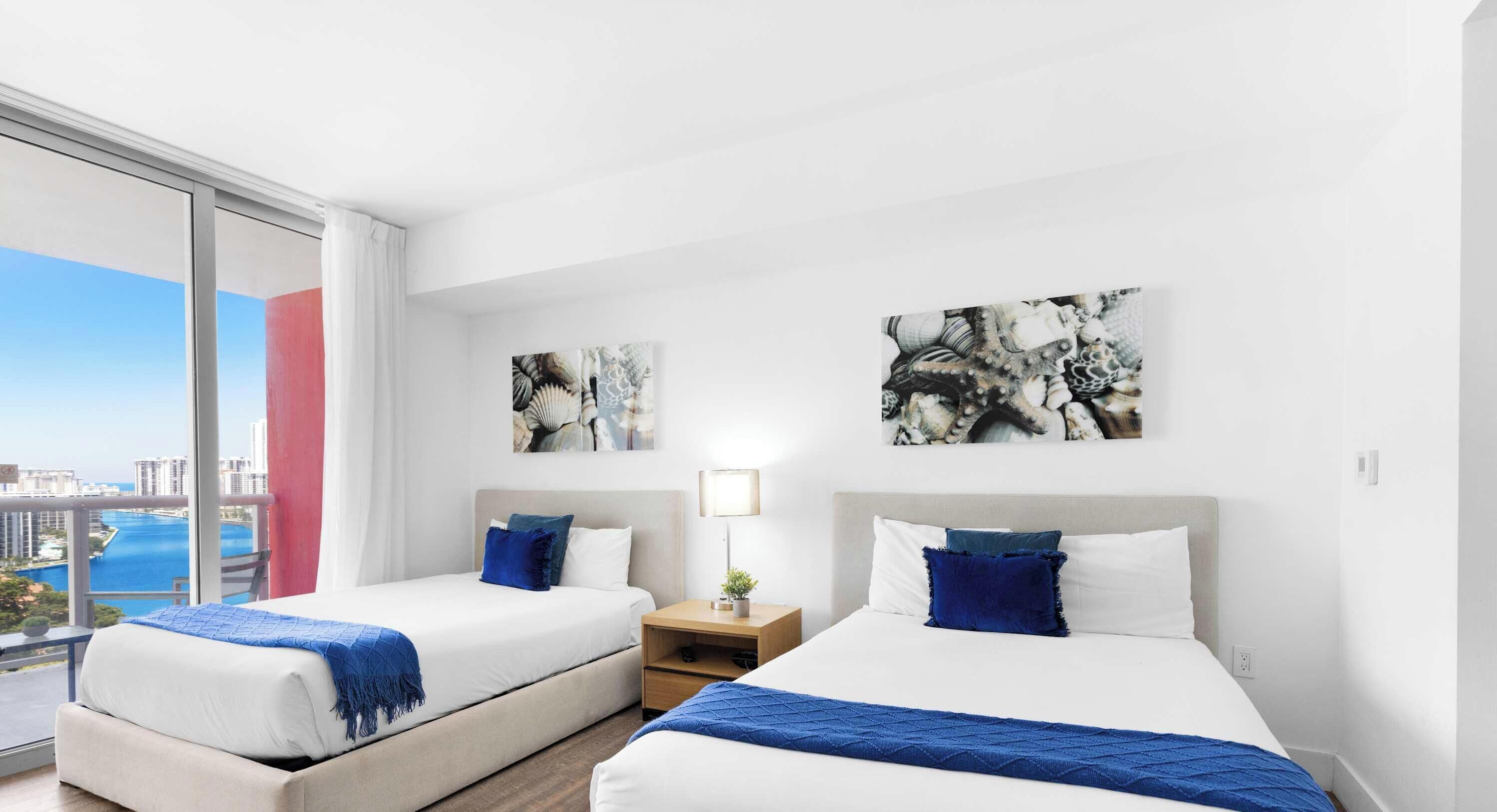 A hotel room with two beds, a nightstand, and a view of the city and water.