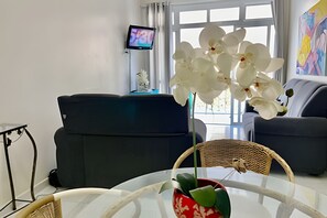 A modern living room with a glass coffee table, a wicker chair, and a sofa. A flat-screen TV is mounted on the wall, and there is a window with white curtains. - SHB - Delicioso apartamento no Guarujá (Guarujá)
