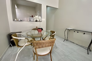 A dining area with a glass table surrounded by wicker chairs, a small red vase, and a white wall with a mirror.