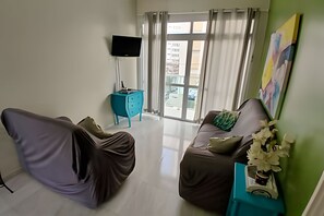 Un salon comprenant un canapé, une table d’appoint de couleur turquoise, un téléviseur à écran plat et une grande fenêtre avec des rideaux.