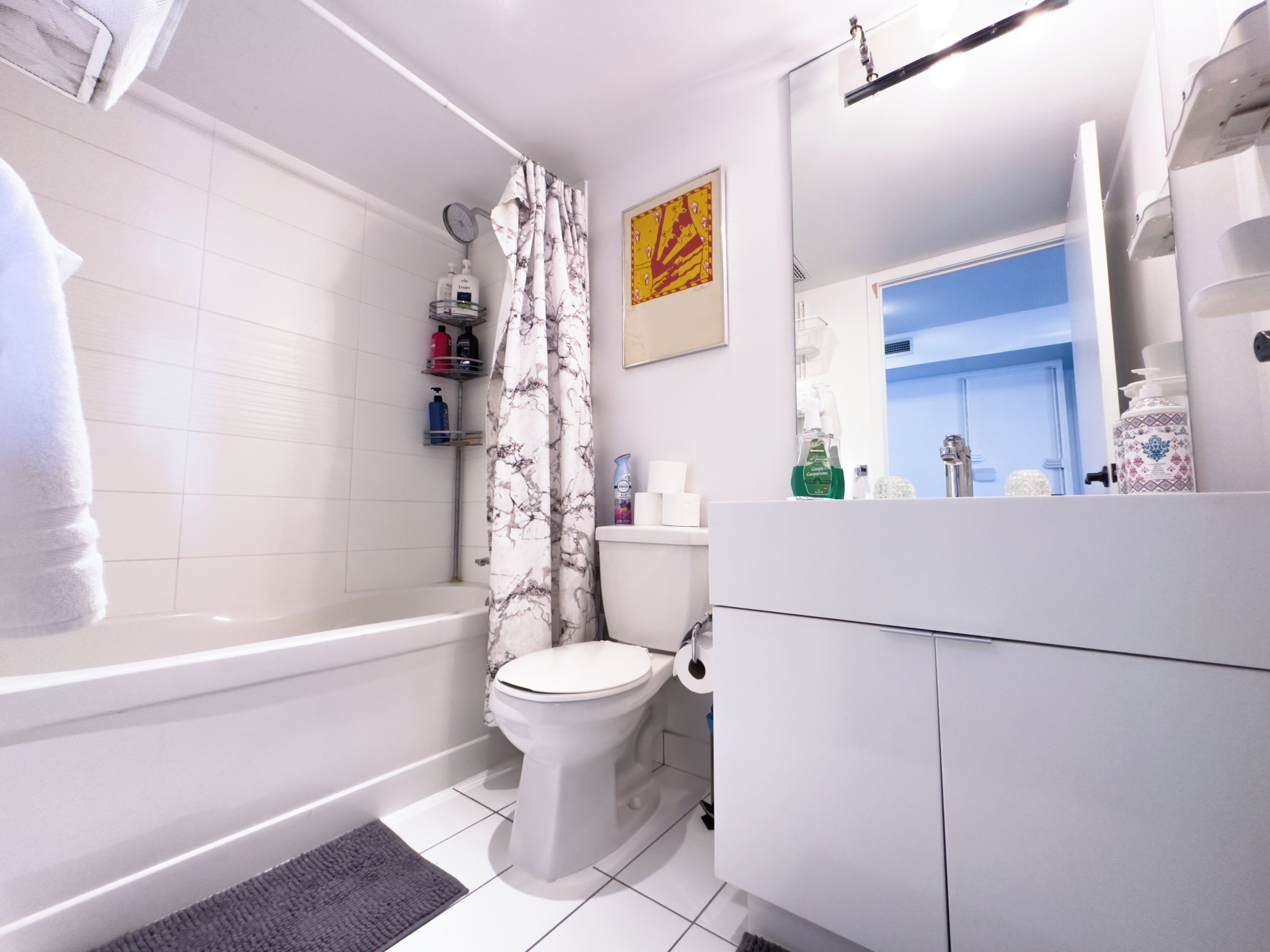 A modern bathroom with a bathtub, toilet, and a shower curtain. There are toiletries on a shelf and a mirror above the sink.