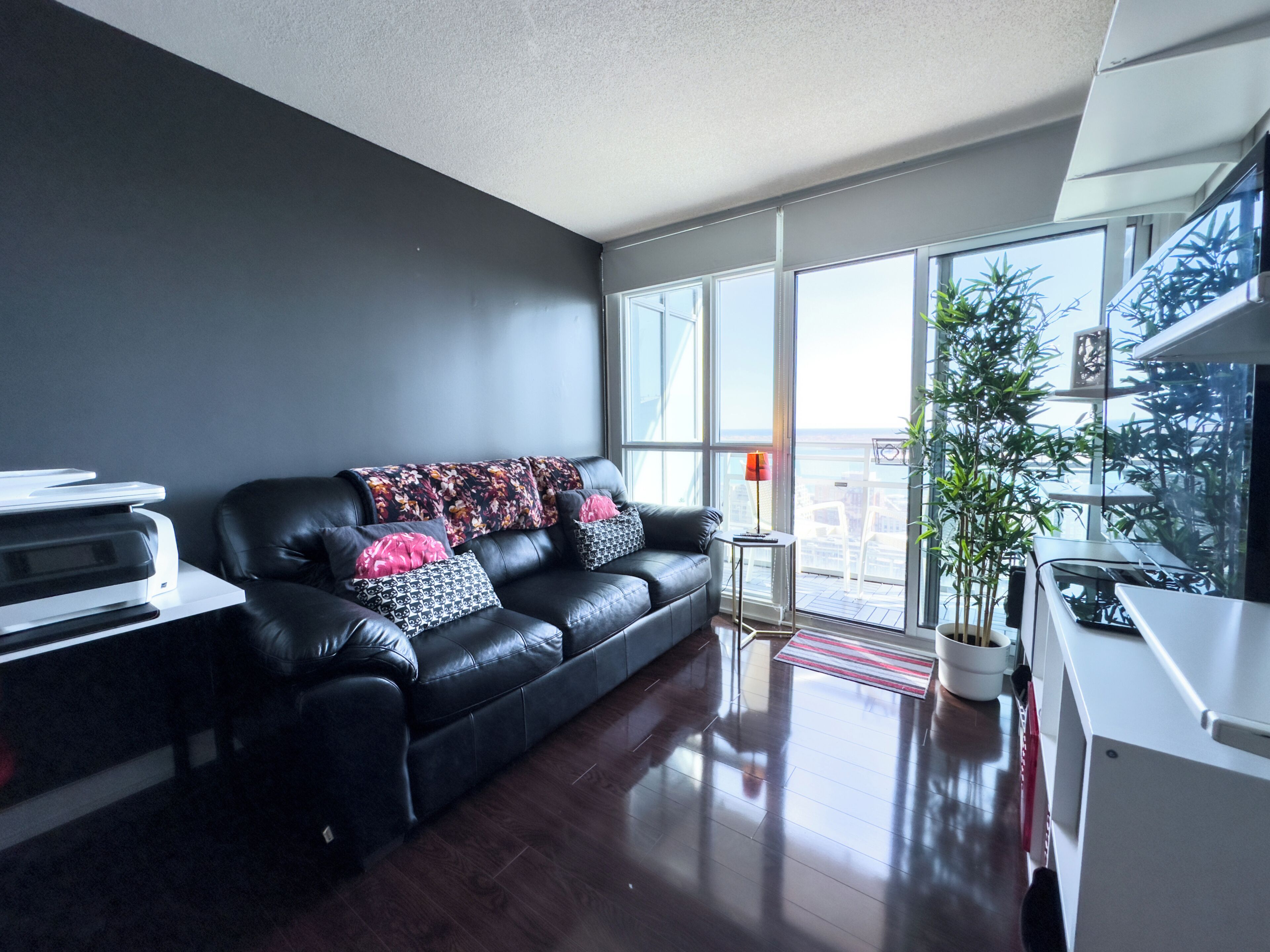 A modern living room with a black leather sofa, a small table with a printer, and a large window offering a view of the city.