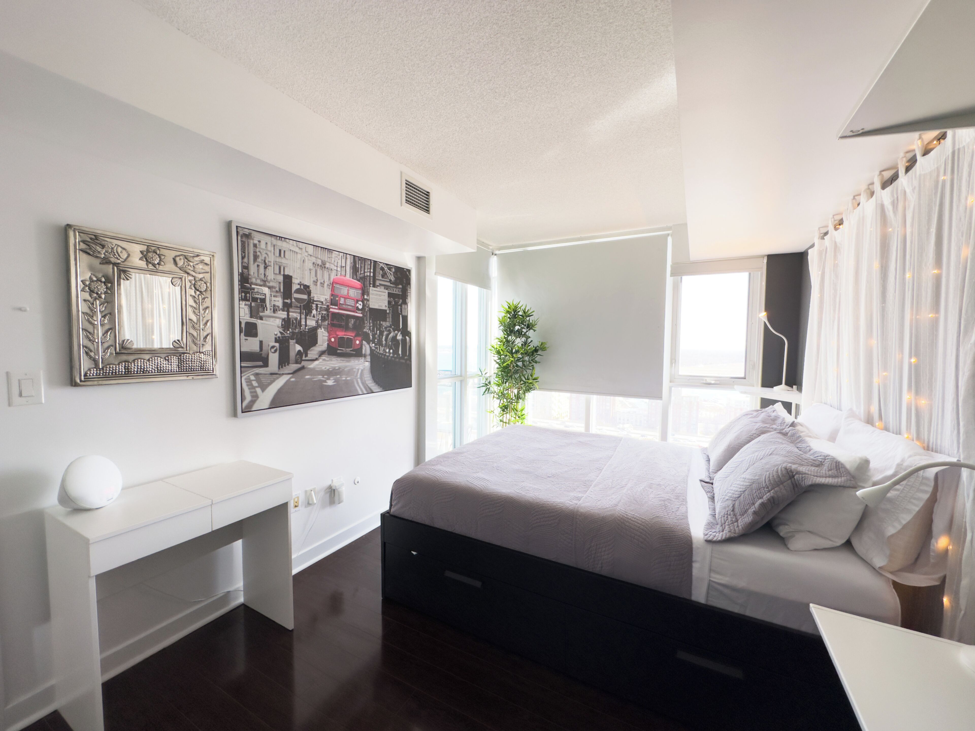 A modern bedroom with a bed, bedside table, and a large window. There are two wall-mounted artworks and a potted plant.
