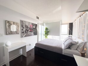 A modern bedroom with a bed, bedside table, and a large window. There are two wall-mounted artworks and a potted plant. - City Central Residences Downtown Toronto (Toronto)