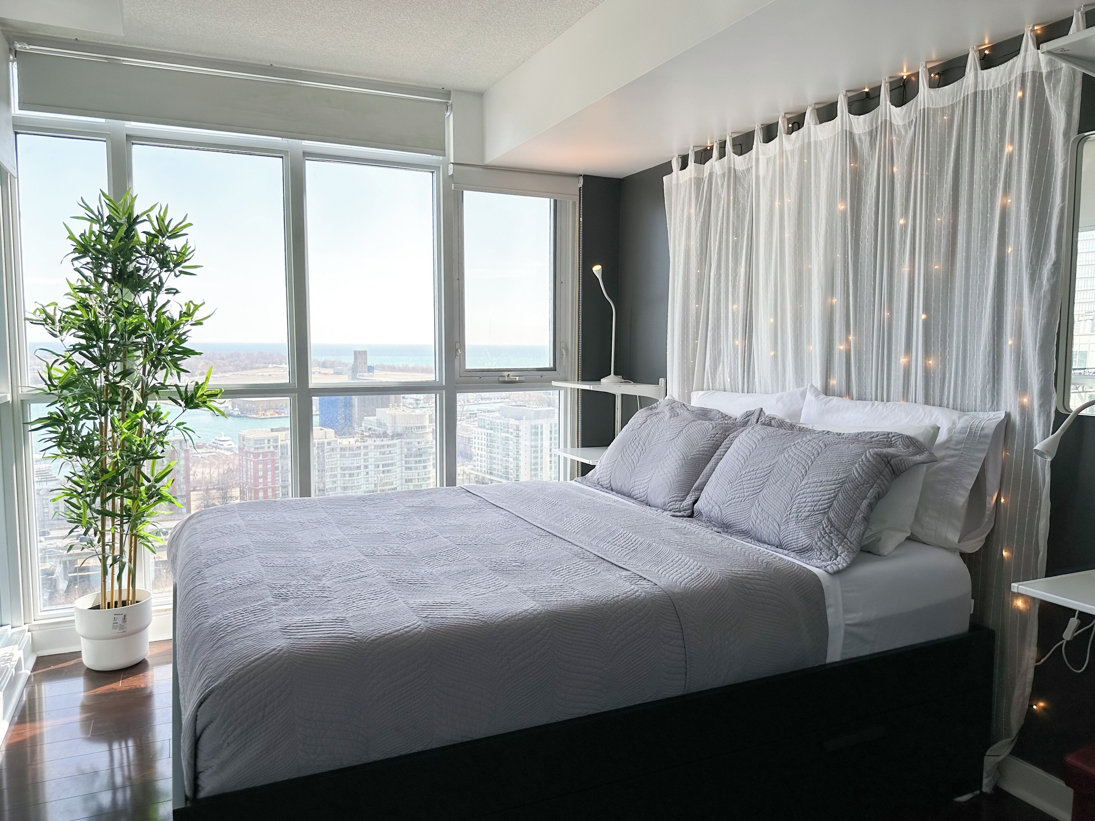 A modern bedroom with a large bed, a potted plant, and a view of the city through large windows.