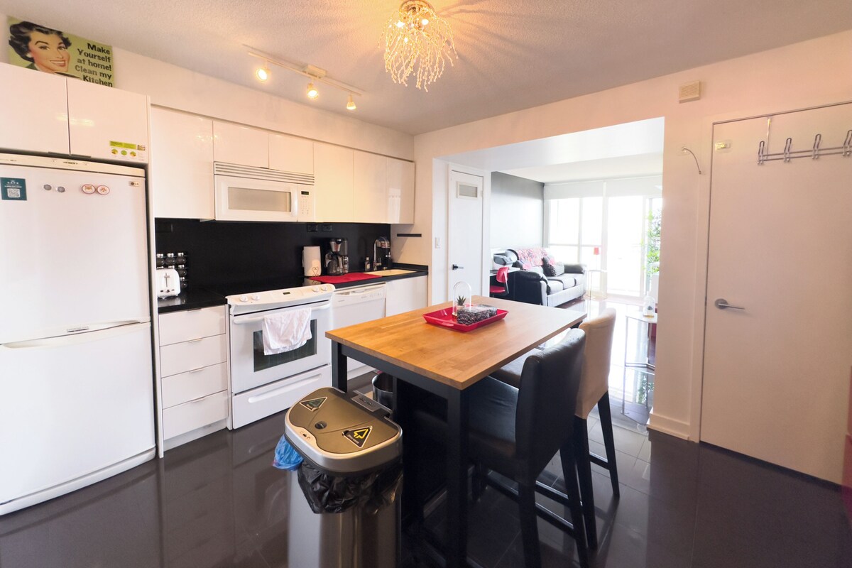 A modern kitchen with white cabinets, a microwave, a stove, and a sink. There is a wooden table with chairs, a trash bin, and a ceiling light fixture.
