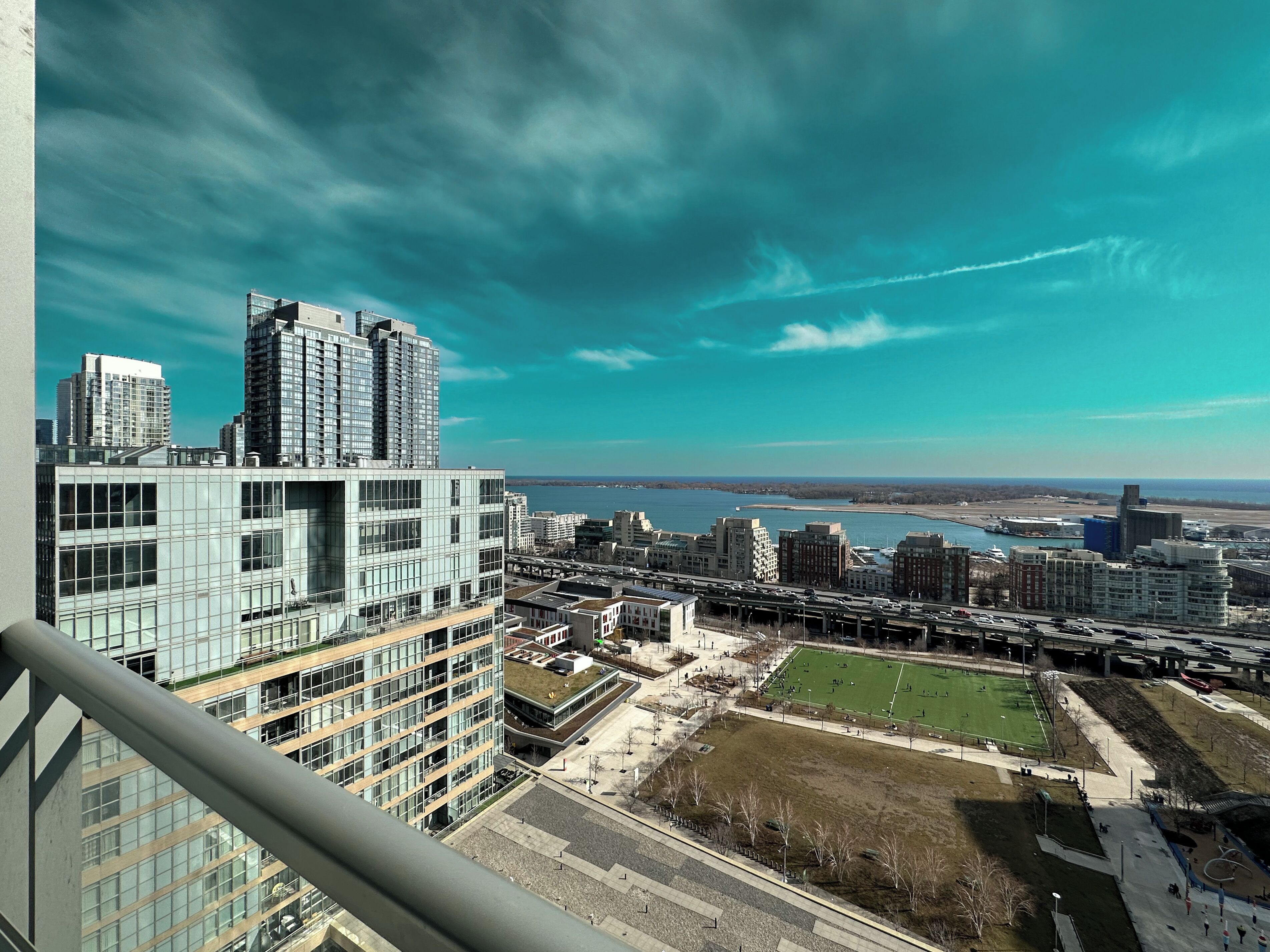 A high-rise building with a modern glass facade, a large open plaza with a green lawn, and a view of a river and distant city skyline.