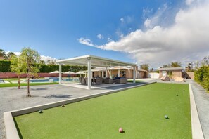 Un área de recreación con un césped de croquet, asientos bajo un pabellón y una piscina al fondo.