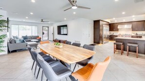 Uma cozinha moderna com armários escuros e uma ilha central, uma sala de jantar com mesa e cadeiras de madeira, e uma sala de estar com sofá e um painel decorativo.