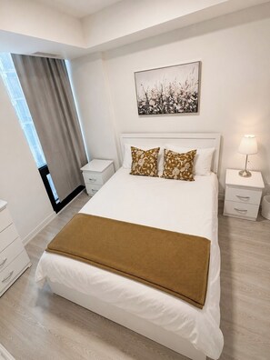 A modern bedroom with a large bed, a bedside table, a nightstand, a lamp, and a window with curtains. - Pleasant 1-bedroom condo -Downtown (Toronto)