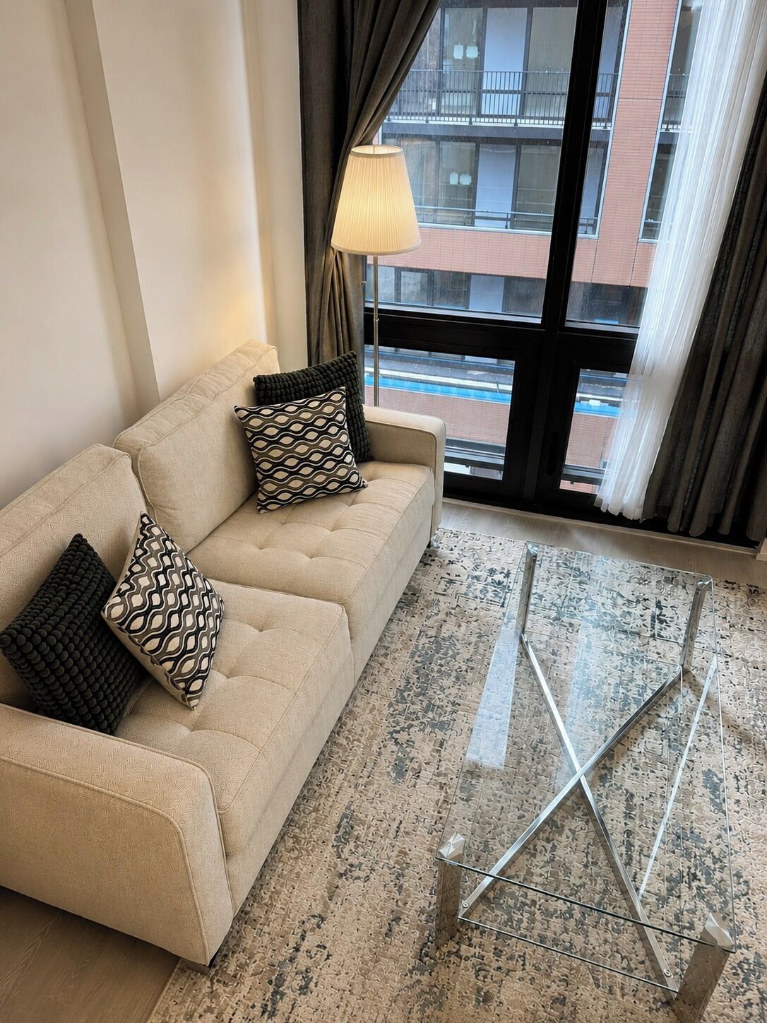 Ein beiges Sofa mit gemusterten Kissen, eine Stehlampe und Blick auf Gebäude durch ein Fenster.