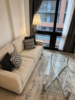 Un sofá color beige con cojines estampados, una lámpara de pie y vistas a edificios a través de una ventana.