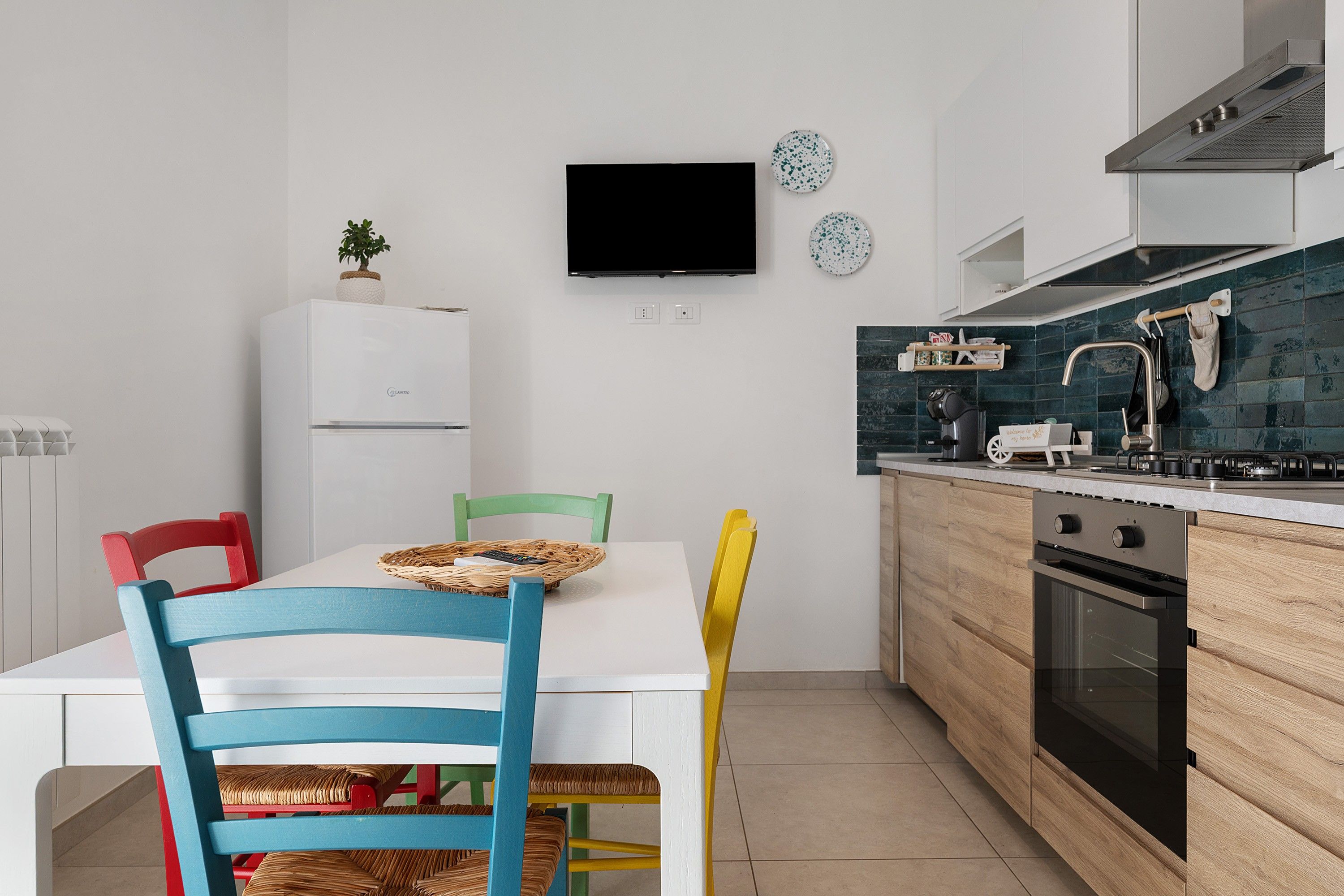 A modern kitchen with a white refrigerator, a built-in oven, and a stove. There is a dining table with colorful chairs and a television mounted on the wall.