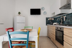 A modern kitchen with a white refrigerator, a built-in oven, and a stove. There is a dining table with colorful chairs and a television mounted on the wall.