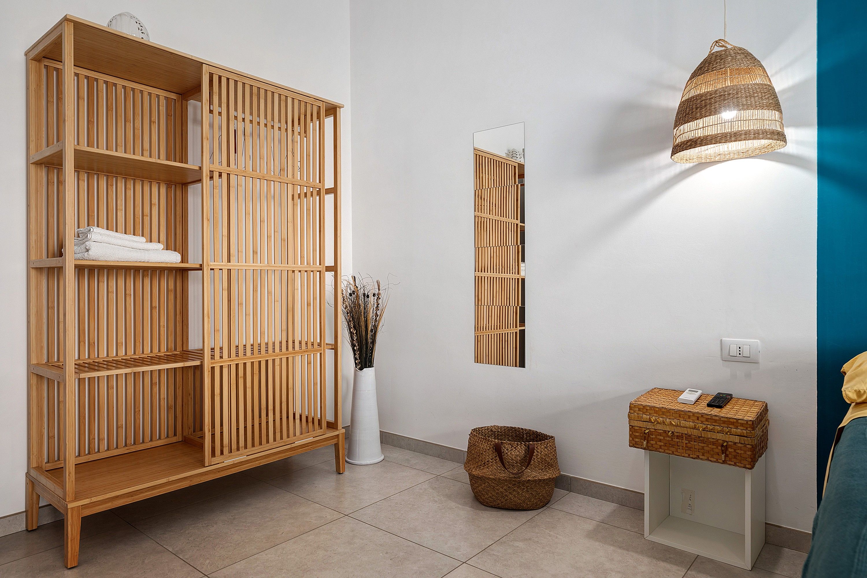 A room with a large wooden bookshelf, a mirror, a wicker lamp, a wicker basket, and a small table with a device.