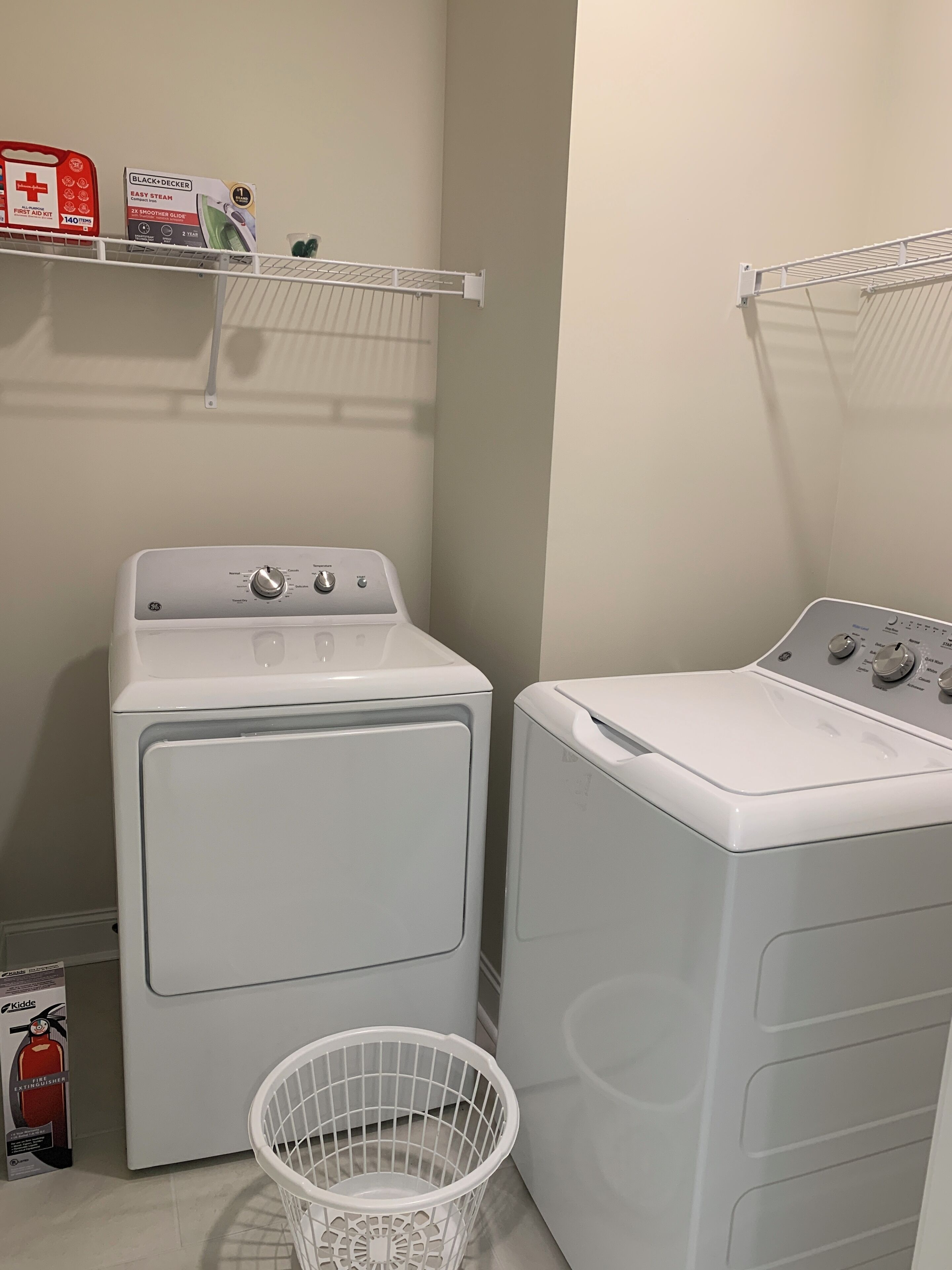 Een wasruimte met twee voorladers wasmachines en drogers, een witte wasmand en een plank met diverse spullen, waaronder een brandblusser.