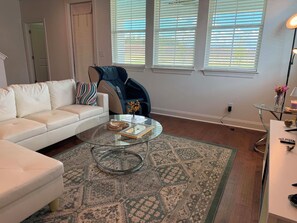 A living room with a white sofa, a glass coffee table, a patterned rug, a blue chair, and a window with blinds. - Cozy Southern Retreat 3 (Oxford)