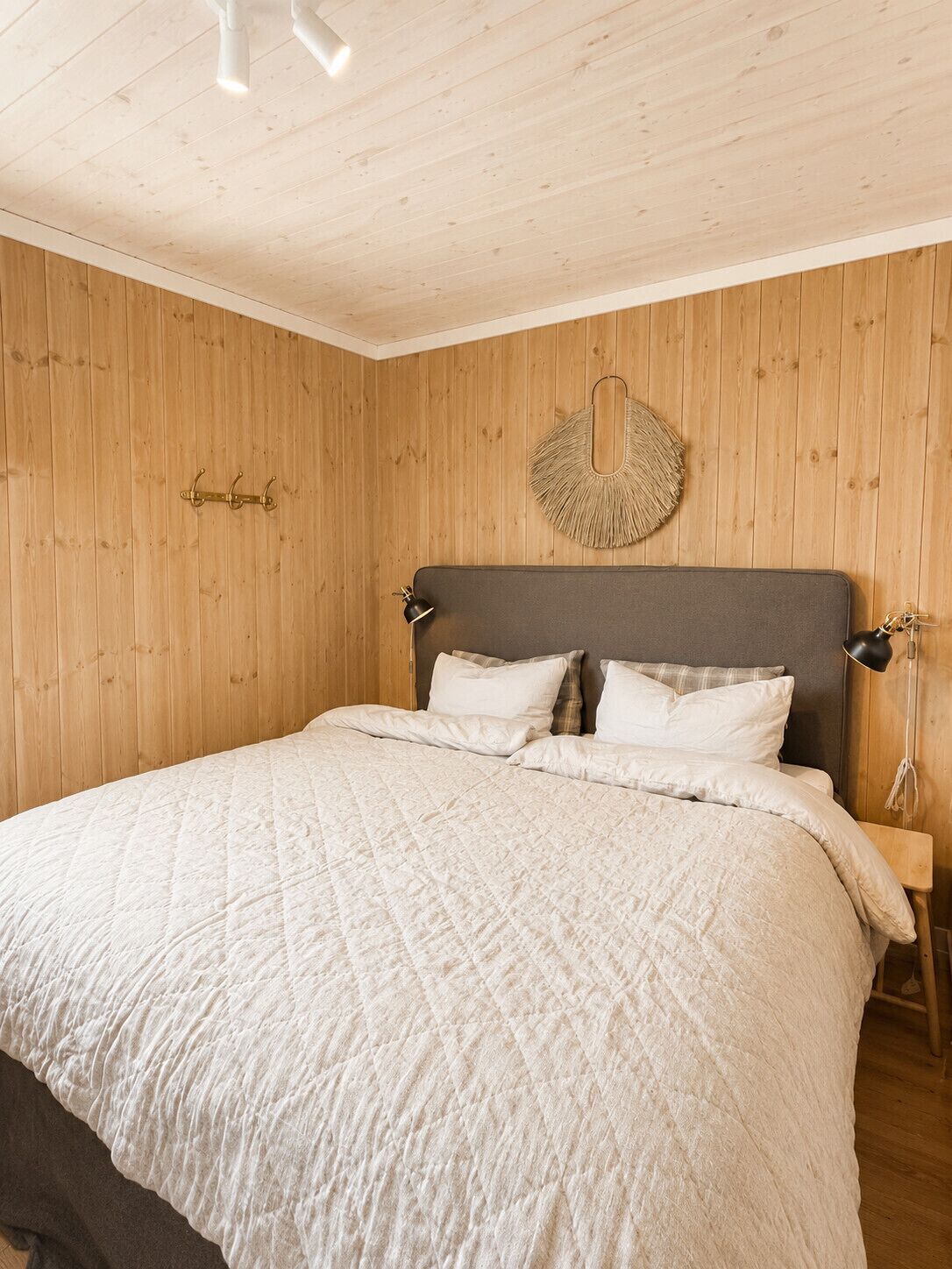 A bedroom with a wooden wall, a bed with a quilted bedspread, two bedside lamps, and a wall-mounted round decoration.
