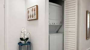 A laundry room with a washing machine and dryer, a blue side table with a potted plant, and a framed picture on the wall. - Comfy Home 4 Beds Steps to Hard Rock Stadium Free Parking Miami Gardens (Miami)