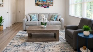 A living room with a grey sofa, a coffee table, and a cityscape painting on the wall. - Comfy Home 4 Beds Steps to Hard Rock Stadium Free Parking Miami Gardens (Miami)