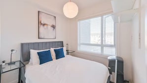 Une chambre à coucher comprenant un lit, une table de chevet, une lampe, une fenêtre avec des stores et un luminaire au plafond.