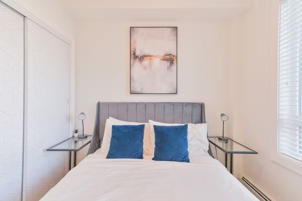 A bedroom with a bed, two bedside tables, a wall-mounted abstract painting, and a window with blinds.