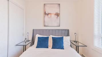 Une chambre à coucher comprenant un lit, deux tables de chevet, un tableau abstrait accroché au mur et une fenêtre avec des stores.