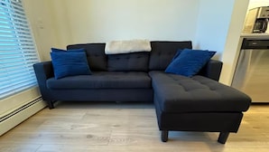 Un canapé d’angle de couleur sombre, agrémenté d’un plaid blanc et de coussins bleus, est placé sur un parquet clair.