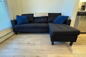 A dark-colored sectional sofa with a white throw blanket and blue cushions, positioned on a light wooden floor.