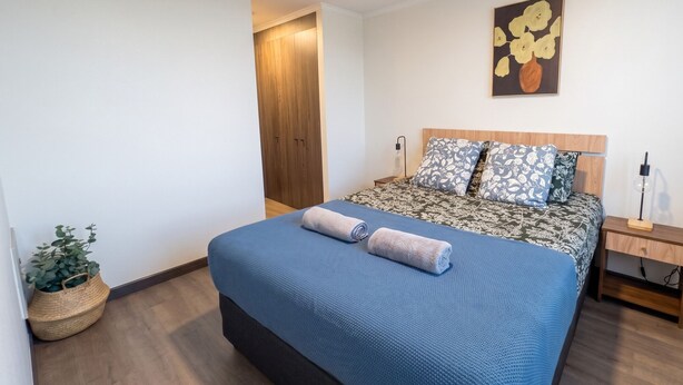 A bedroom with a bed, a nightstand, a lamp, a wall art piece, and a potted plant. - Parque Los Reyes - Domus Travels (Santiago)