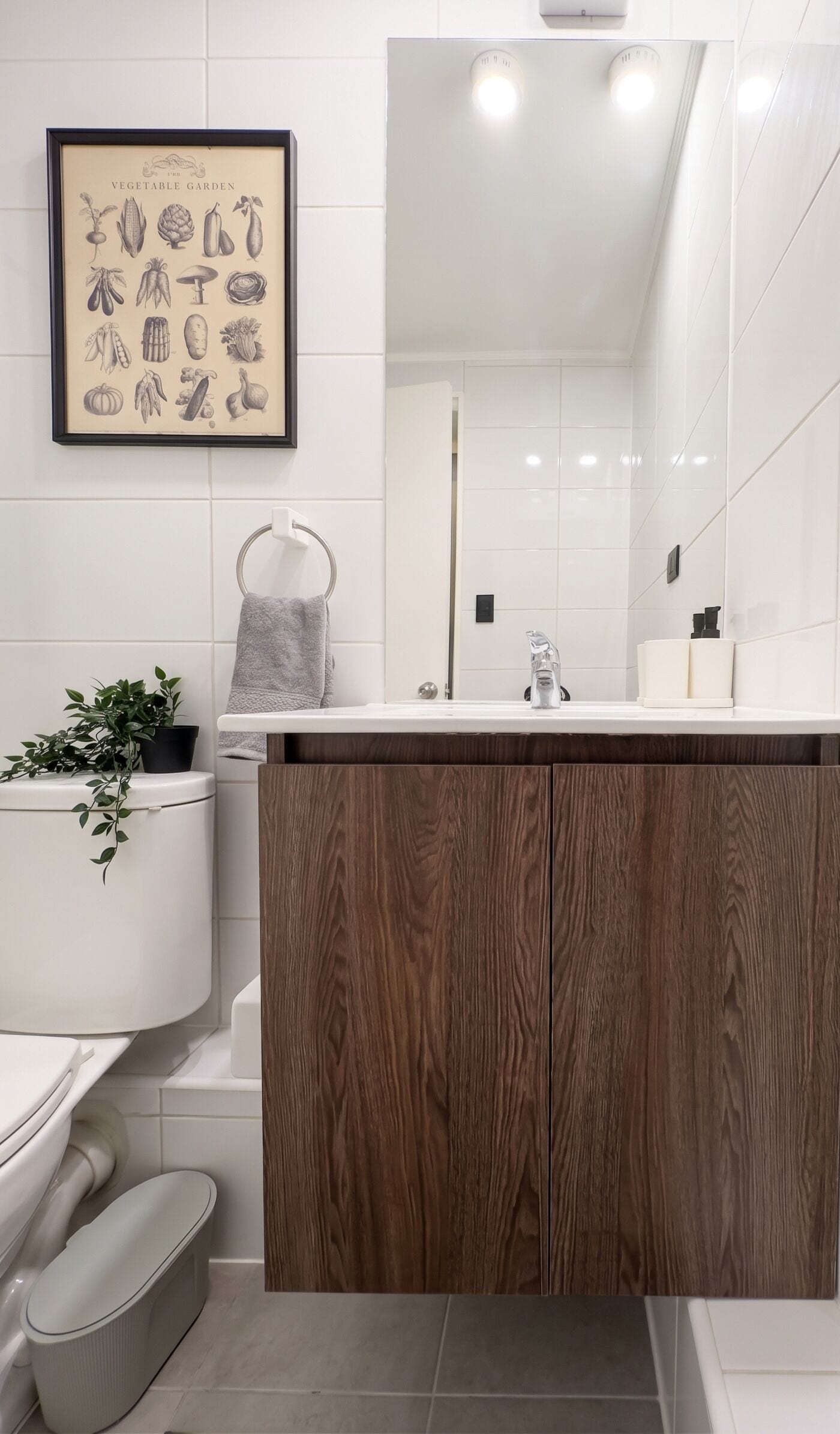 A modern bathroom with a wooden vanity, a framed botanical illustration, and a white tiled wall.