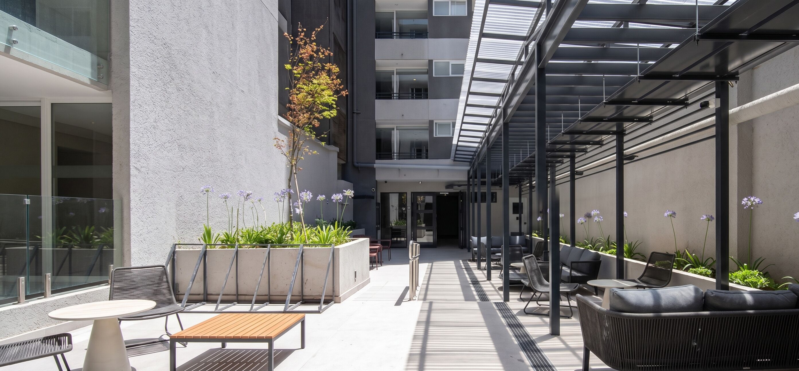 A modern outdoor seating area with tables and chairs, surrounded by plants and a glass railing.