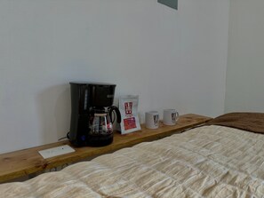 A bed with a white quilt, a wooden bedside table with a black coffee maker, a packet of coffee, and two white mugs.