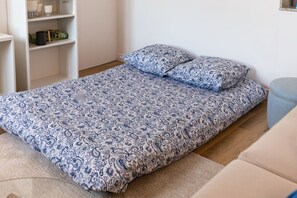 Un llit individual amb un llençol estampat i dos coixins, una petita taula de nit i un sofà de color clar en una habitació amb parets blanques i un terra de fusta.