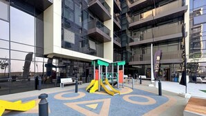An outdoor play area with a slide and climbing structures, surrounded by a building with large windows. - Brand-New 1BR in Neva Residence | Near Circle Mall (Dubai)